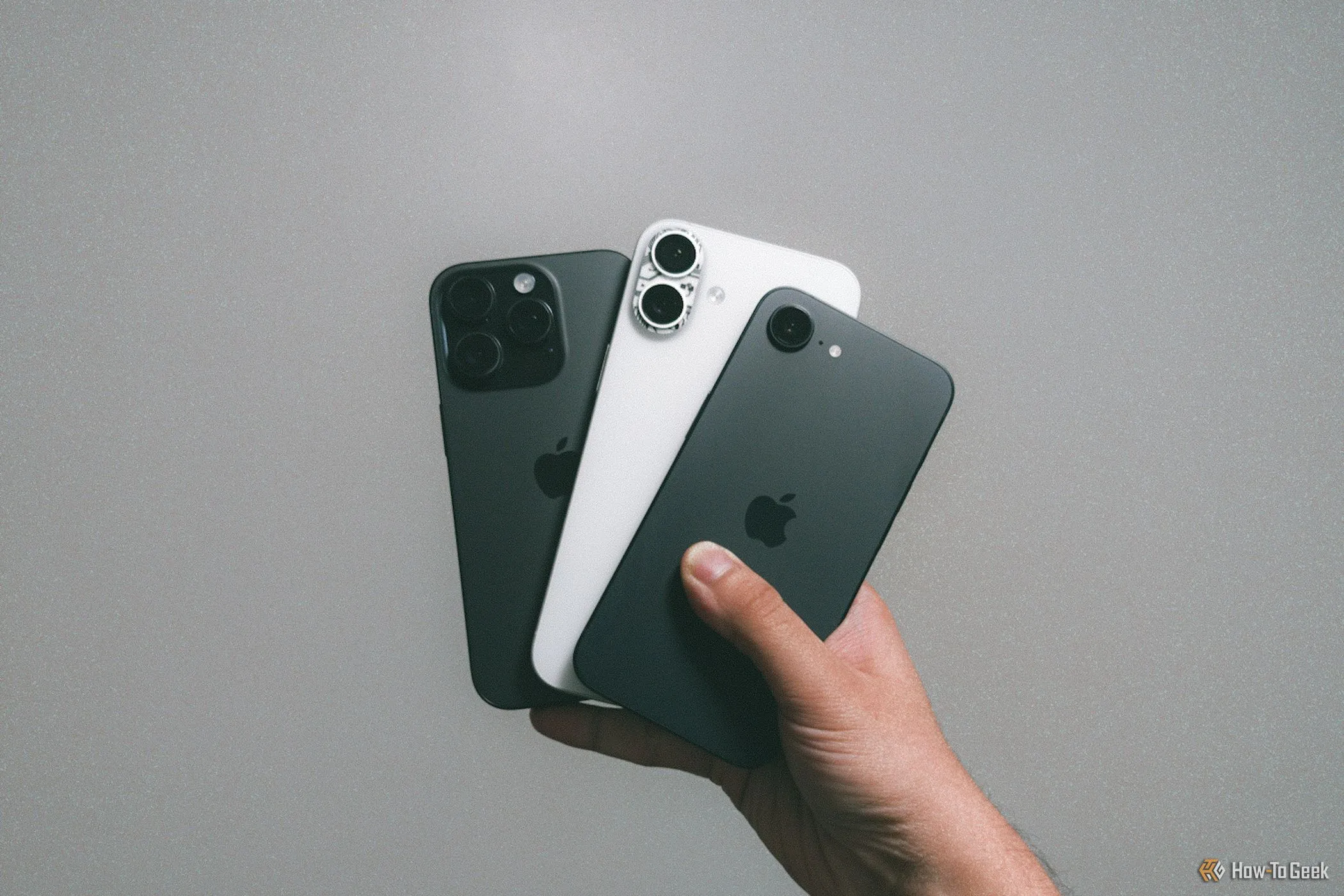 Ba mẫu iPhone 16e, iPhone 16 Plus và iPhone 16 Pro được cầm trên tay dựa vào bức tường xám, thể hiện sự khác biệt về kích thước và tính năng giữa các phiên bản.