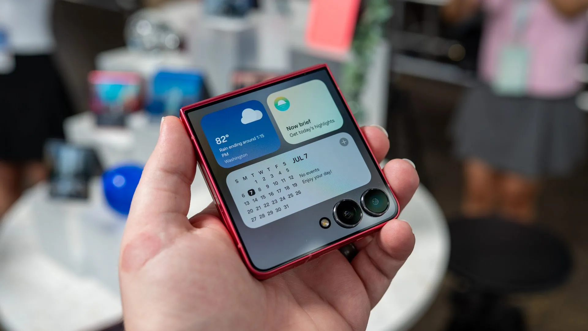 Bàn tay đang cầm điện thoại Samsung Galaxy Z Flip 7, hiển thị các widget thông báo trên màn hình phụ FlexWindow, giúp người dùng quản lý thông tin nhanh chóng mà không cần mở máy.