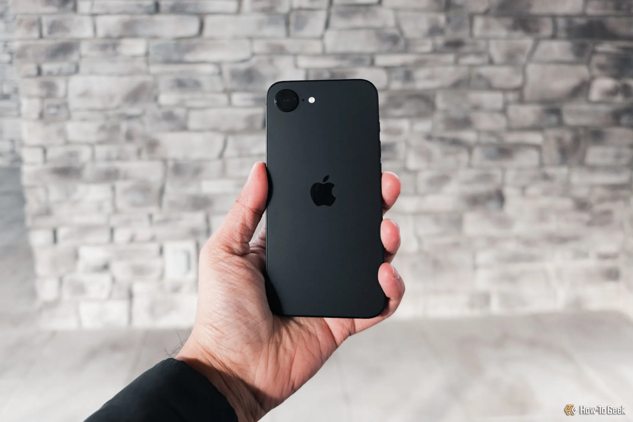 iPhone 16e màu đen được cầm trên tay dựa vào bức tường xám, thể hiện khả năng sử dụng bền bỉ cả ngày nhờ thời lượng pin được cải thiện đáng kể.