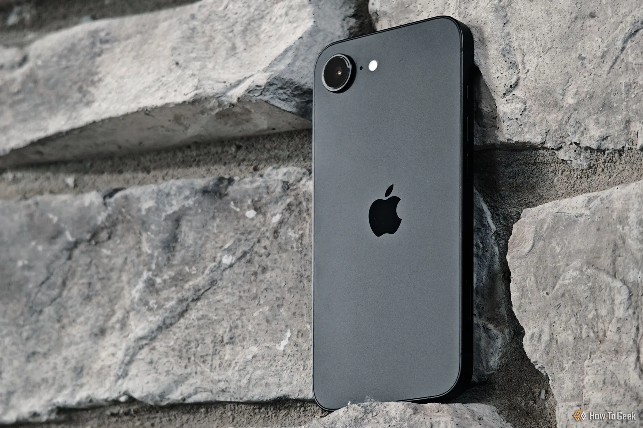 iPhone 16e màu đen tựa vào bức tường gạch, minh họa thiết kế tổng thể của điện thoại nhưng không hỗ trợ sạc MagSafe.