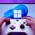 Một người đang cầm tay cầm Xbox One màu đen, phía sau là màn hình máy tính với hình nền Windows 11.